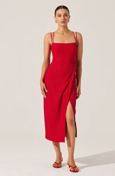 A woman is wearing a long, fitted red dress with a textured fabric. The dress has thin double spaghetti straps over her shoulders, a straight neckline, and a side slit that reveals her leg. She is also wearing red high-heeled sandals with thin straps.